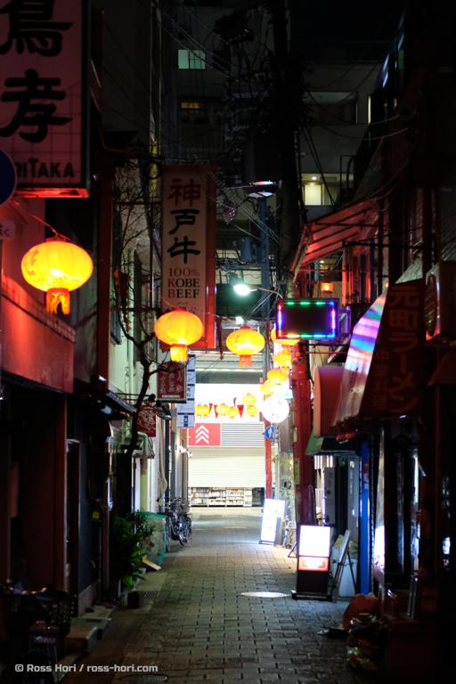 An alleyway in Kobe, Japan. The darkness is lit with Chinese lanterns and the glow of signs calling people to go inside.