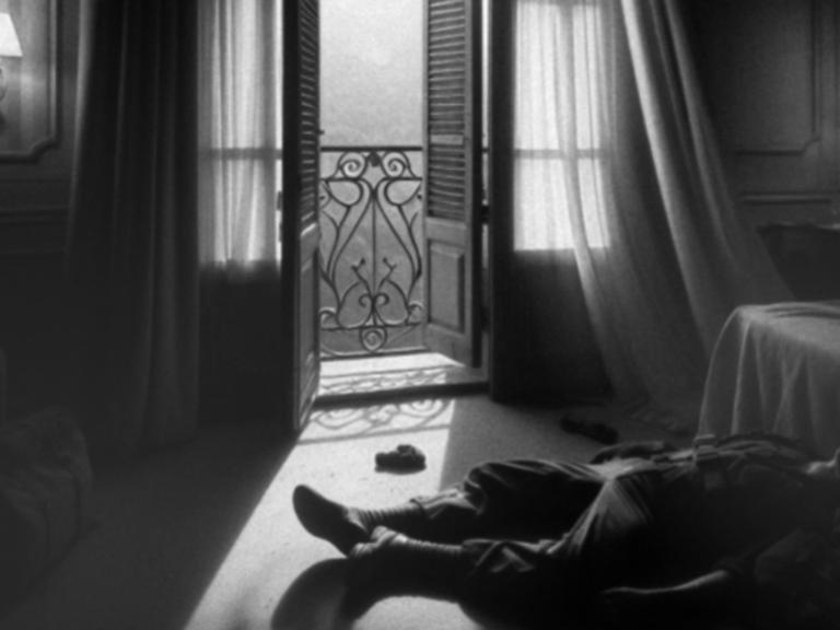 Black and white image of a paratrooper lying on the floor of a hotel room with the balcony doors open and a wind wafting through net curtains.